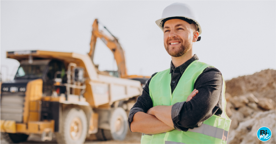 A construction worker smiling with his arms folded and a big construction vehicle in the background